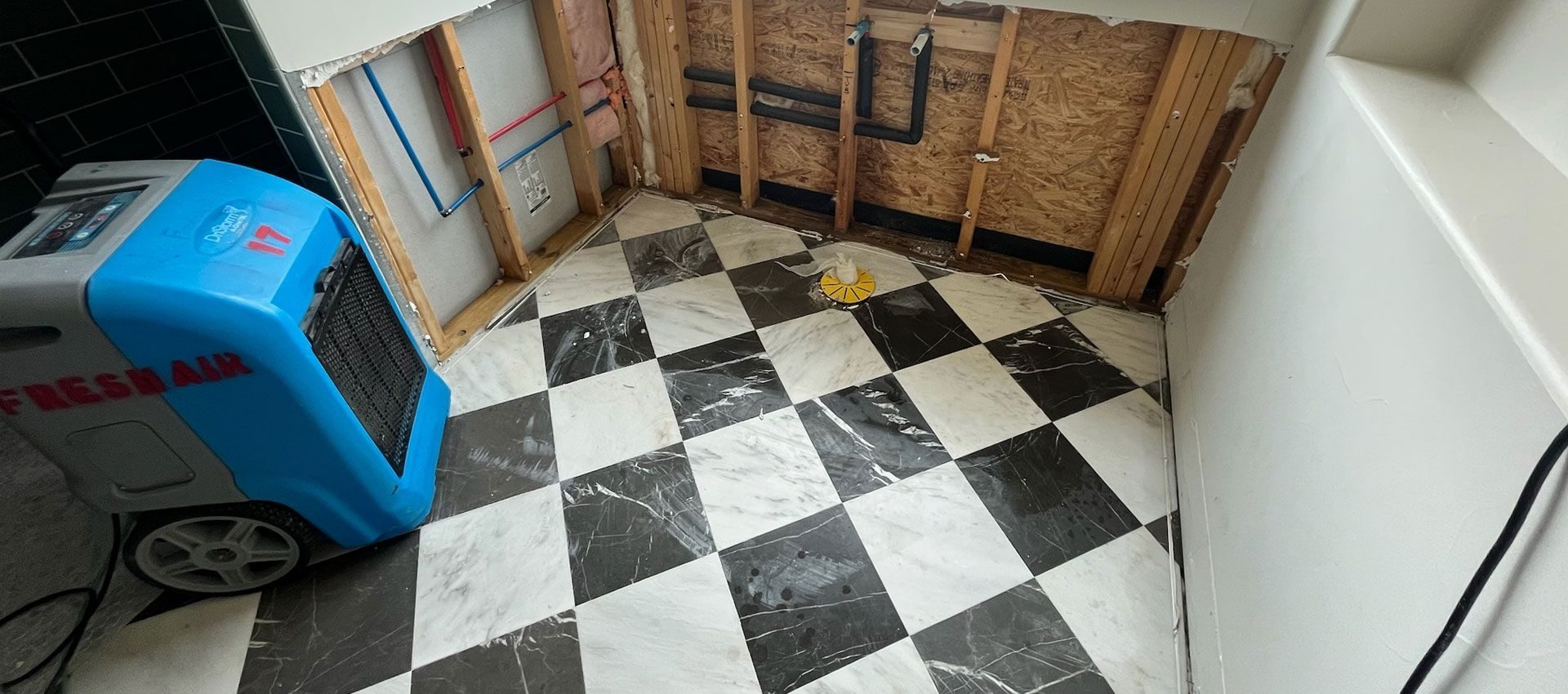 Water-damaged bathroom with walls opened to the studs and a dehumidifier running on the checkered tile floor