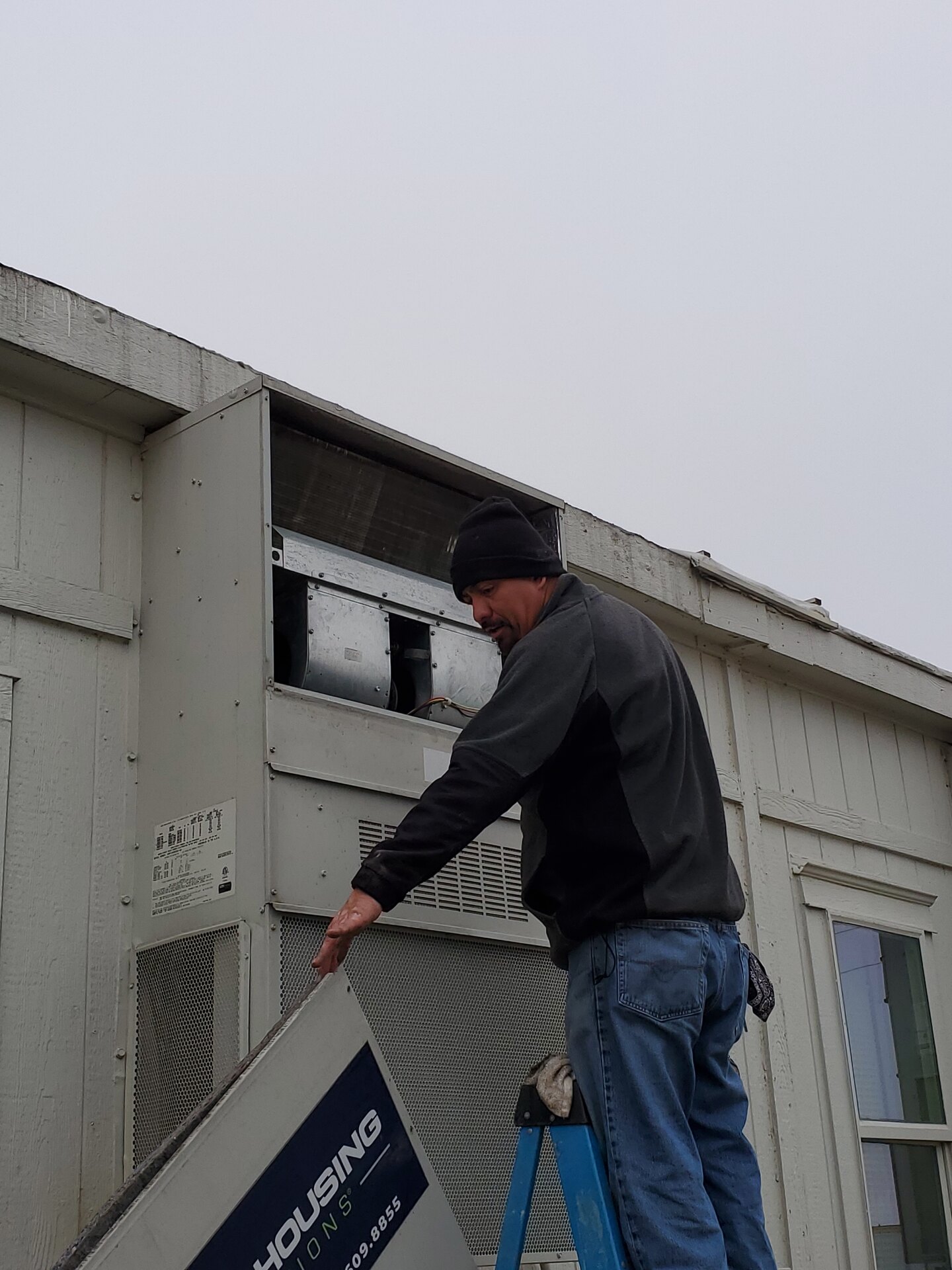 Fresh Air technician on a ladder removing the access panel from a wall-mounted commercial HVAC unit